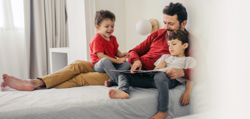 Father sitting on a bed reading a book with his two young sons, all smiling and enjoying their time together in a bright, cozy bedroom.