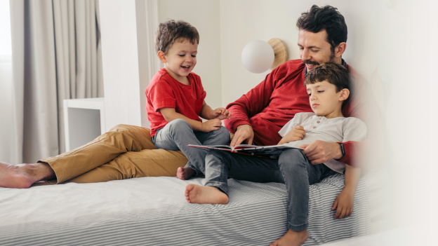 Father sitting on a bed reading a book with his two young sons, all smiling and enjoying their time together in a bright, cozy bedroom.